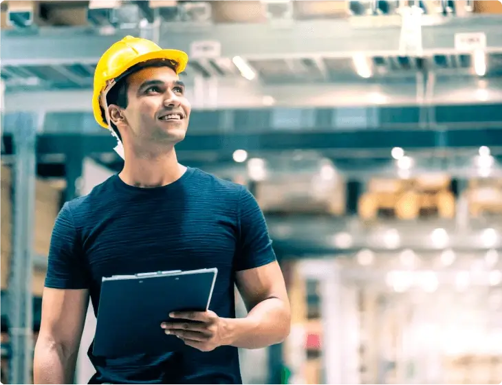 Engenheiro industrial masculino com capacete de segurança amarelo, segurando um tablet e
                 olhando para cima com expressão pensativa em uma fábrica.